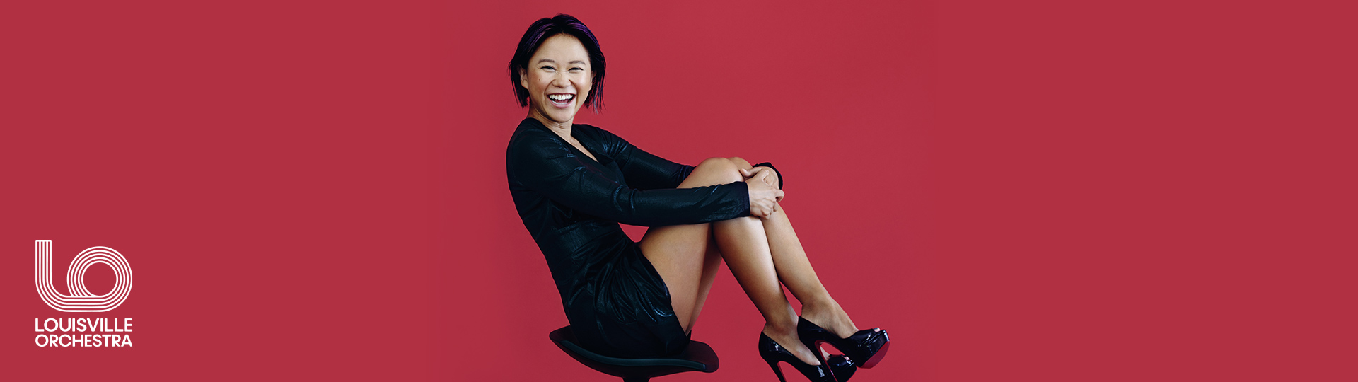 Pianist Yuja Wang wearing a black dress and high heels sitting sideways on a modern chair against a red background.