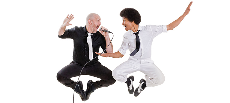 Two men in a mid-air jump against a white background, with the man on the left wearing a black shirt and pants with a white tie and the man on the right wearing a white shirt and pants with a black tie. The man in black is singing into a microphone while the man in white has his arms outstretched.