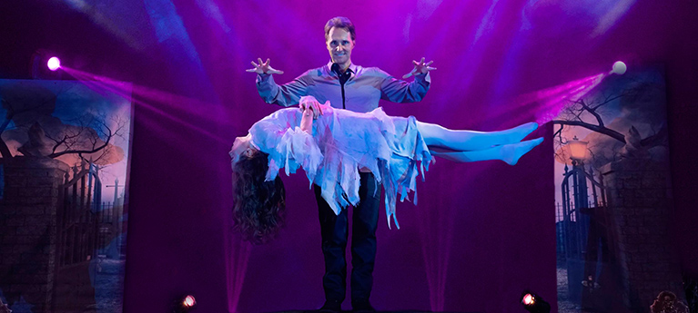Magician levitates a woman in a ghostly white costume on a spooky stage with purple lighting, fog, skulls, and graveyard scenery.