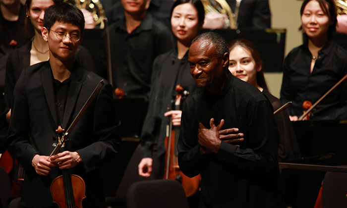 A group of musicians in black attire stands on stage, with one person gesturing with their hand over their heart, during a performance.