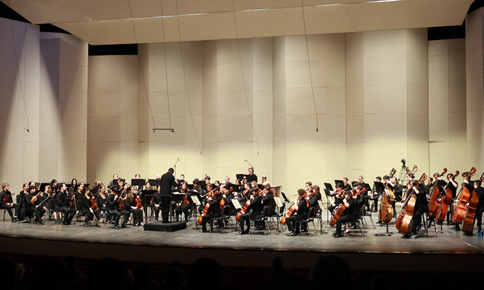 A large orchestra performs on stage, with musicians in black attire and a conductor directing in front of a modern backdrop.