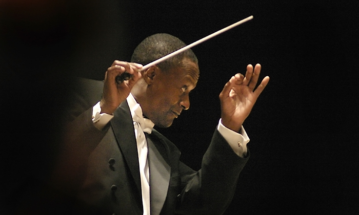 A conductor - Thomas Wilkins - in formal attire gesturing passionately with a baton, leading an orchestra performance against a dark backdrop.