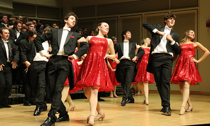 A choreographed performance featuring dancers in sparkly red dresses and tuxedos on stage, with an audience visible in the background.
