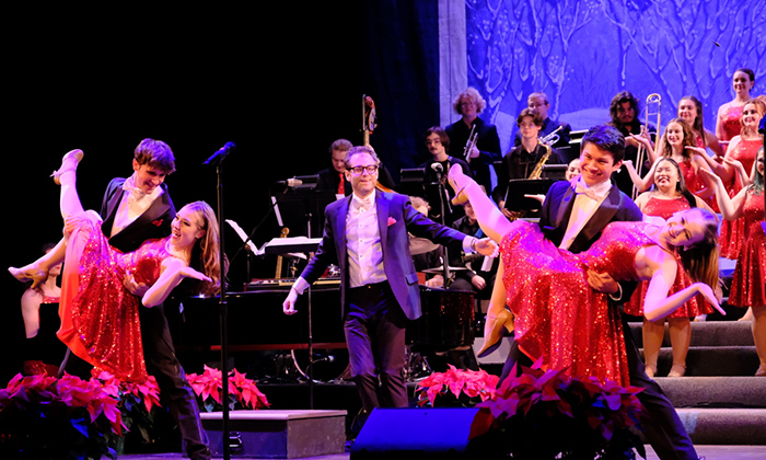 A lively performance with dancers in red sequined dresses and tuxedos, set against an orchestra and festive decorations.