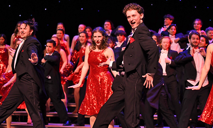 A lively choir dressed in formal tuxedos and sparkling red dresses performs energetically on stage, conveying joy and excitement in a vibrant atmosphere.