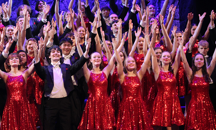 A large group of performers in sparkling red dresses and black suits, joyfully raising their arms on stage.