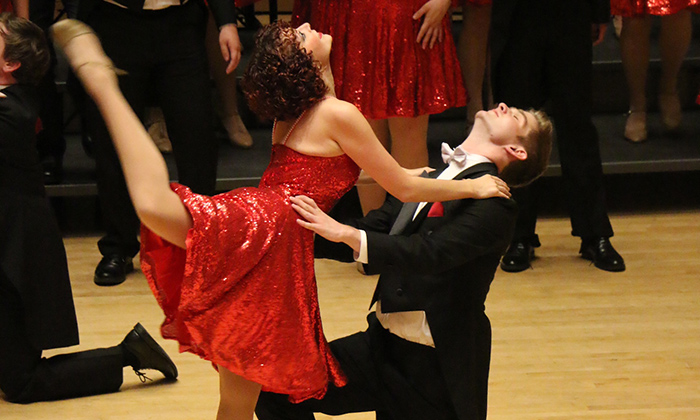 A dancer in a sparkling red dress performs a dramatic pose with a partner in formal attire, showcasing a lively performance on stage.