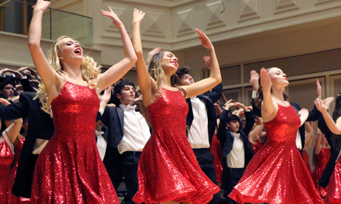 A group of dancers in vibrant red sequined dresses perform energetically on stage. Their raised arms and smiling faces convey joy and excitement.