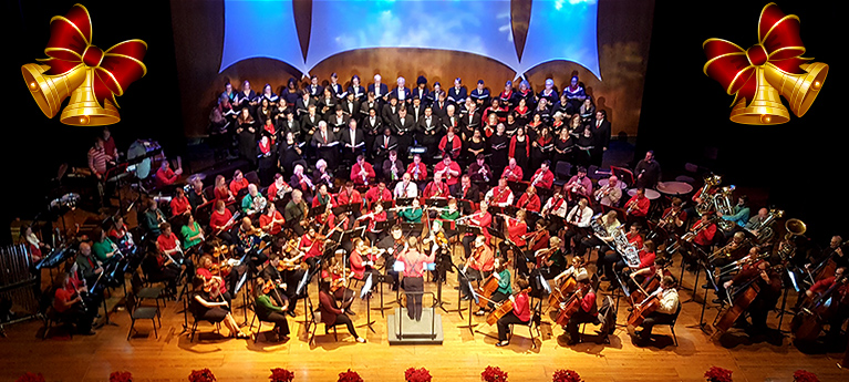 Holiday orchestra and choir performing onstage with Christmas trees, bells, and festive decorations.