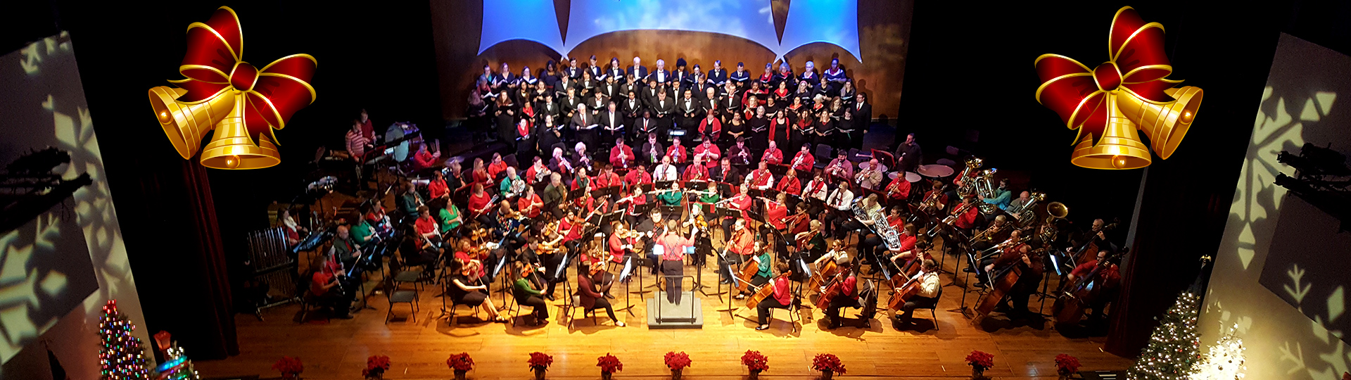 Holiday orchestra and choir performing onstage with Christmas trees, bells, and festive decorations.