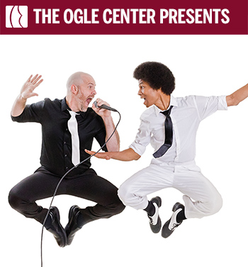 Two men in a mid-air jump against a white background, with the man on the left wearing a black shirt and pants with a white tie and the man on the right wearing a white shirt and pants with a black tie. The man in black is singing into a microphone while the man in white has his arms outstretched.